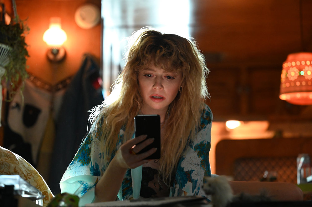 Charlie wears a white and green satin robe while sitting on her bed in her trailer and looking at her phone in Poker Face Season 1 Episode 1, "Dead Man's Hand."