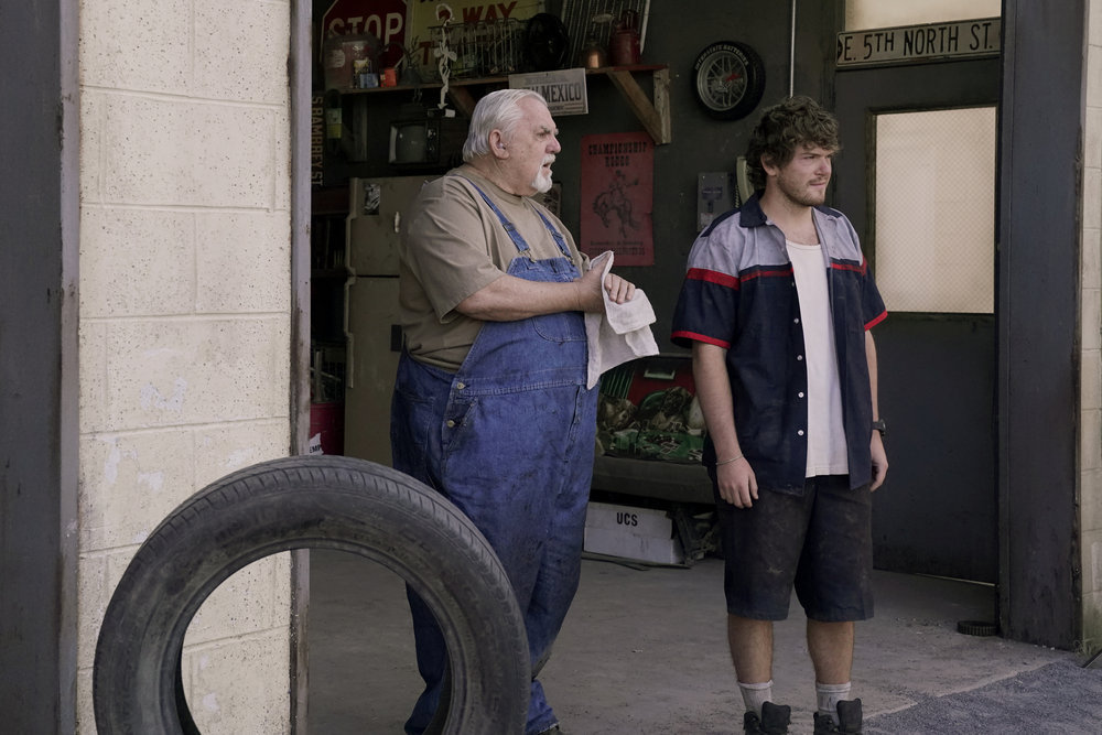 Abe and Jed stand in the open garage of the mechanic shop on a sunny day in Poker Face Season 1 Episode 2, "The Night Shift."