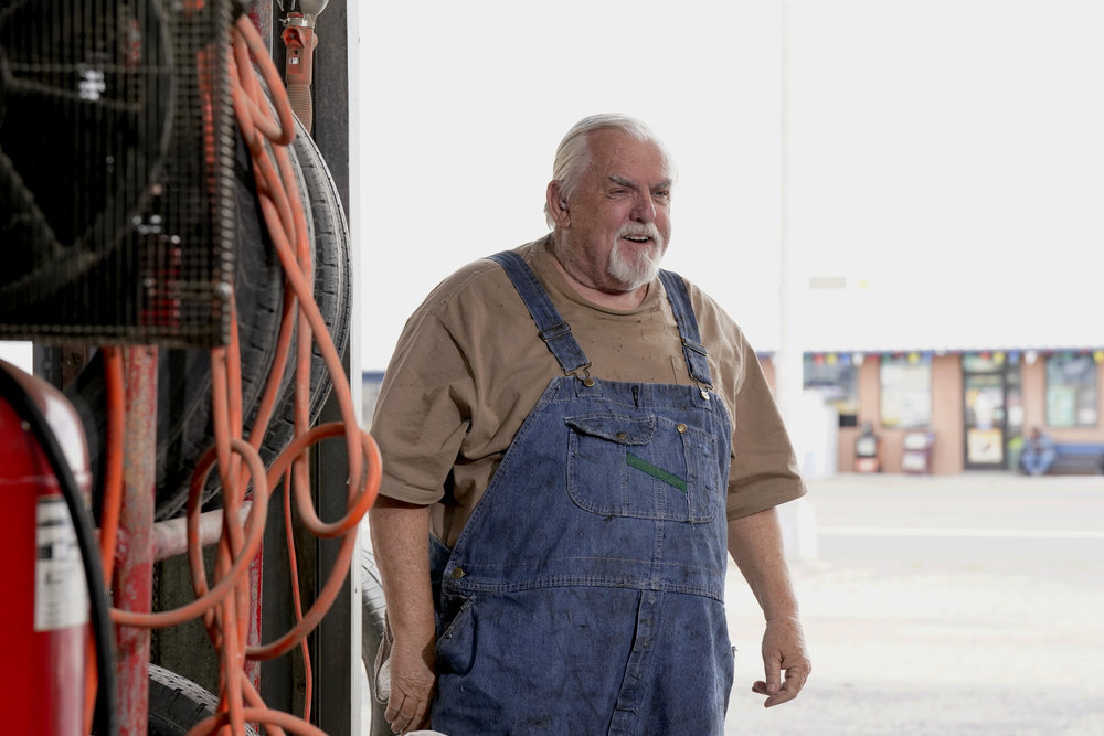 Abe stands in his mechanic shop on a sunny day while wearing denim overalls and a tan shirt in Poker Face Season 1 Episode 2, "The Night Shift."