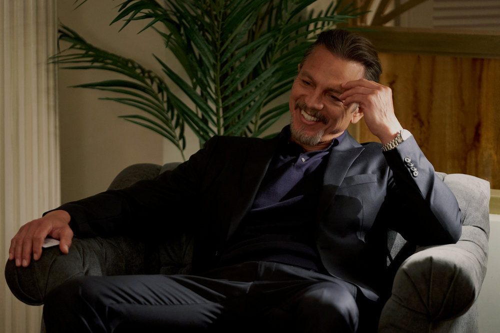 Cliff Legrand sits in a chair in a bedroom suite with his hand on his forehead while laughing in Poker Face Season 1 Episode 1, "Dead Man's Hand."