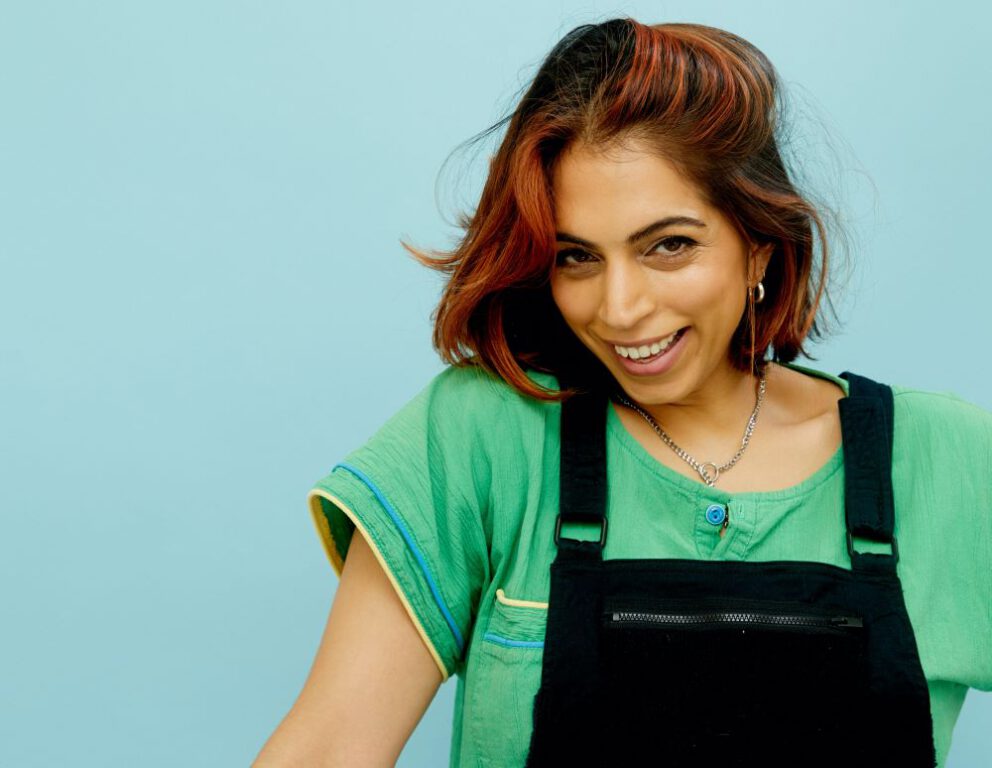 Run the Burbs star Rakhee Morzaria smiles in front of a light blue background while wearing a green short-sleeved shirt and black overalls.