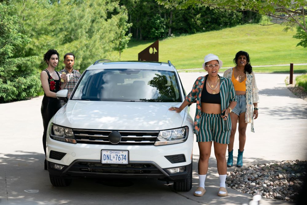 Heath V. Salazar plays the character Arrow in the TV series Sort Of. They stand outside next to a car while wearing a black tank top with red roses and their hair in a ponytail.