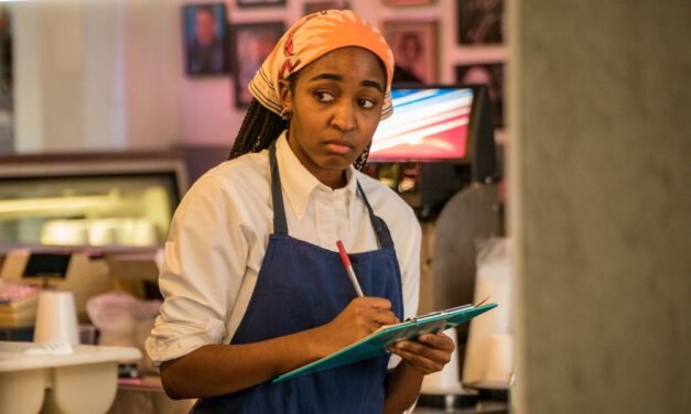 Sydney Adamu, played by Ayo Edebiri, wearing an apron and white shirt while holding a clipboard and pen and standing in a restaurant kitchen in the series The Bear.
