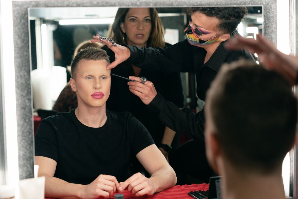 A drag performer sits in a chair while a makeup artist stands next to them and applies makeup in the reality TV show Drag Heals.