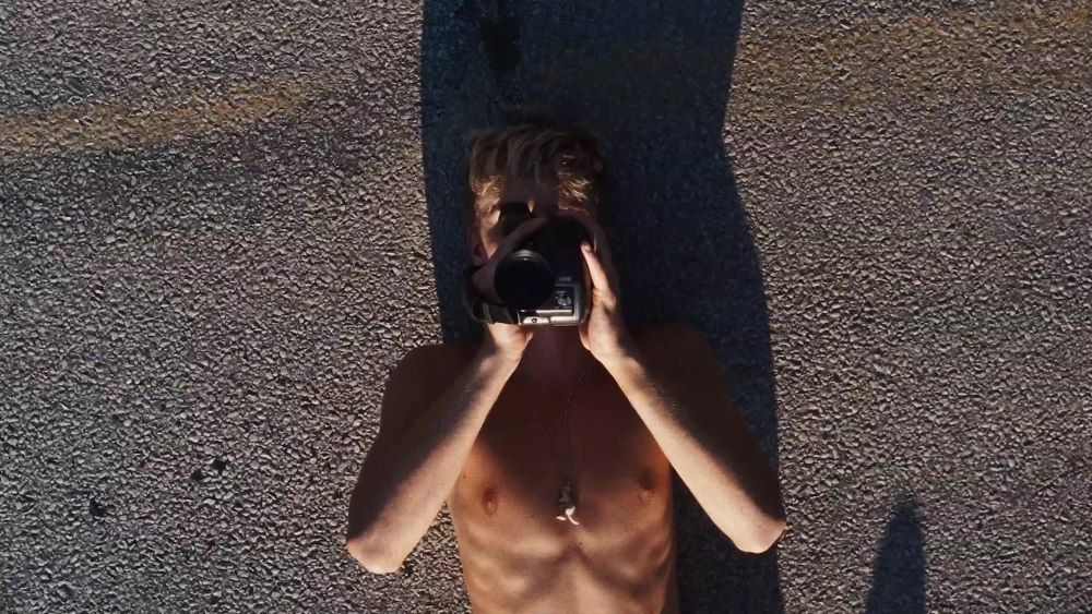 Finn lies on the road shirtless while holding a camera in front of his face in the film American Cherry.