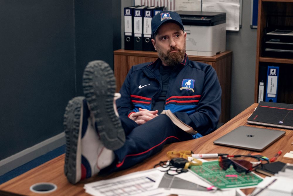 Coach Beard props his legs up on his desk in his office in Ted Lasso Season 3 Episode 1, "Smells Like Mean Spirit."