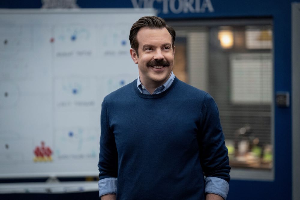 Ted stands in the locker room at AFC Richmond while smiling in front of a dry-erase board in Ted Lasso Season 3 Episode 2, "(I Don't Want to Go to) Chelsea."