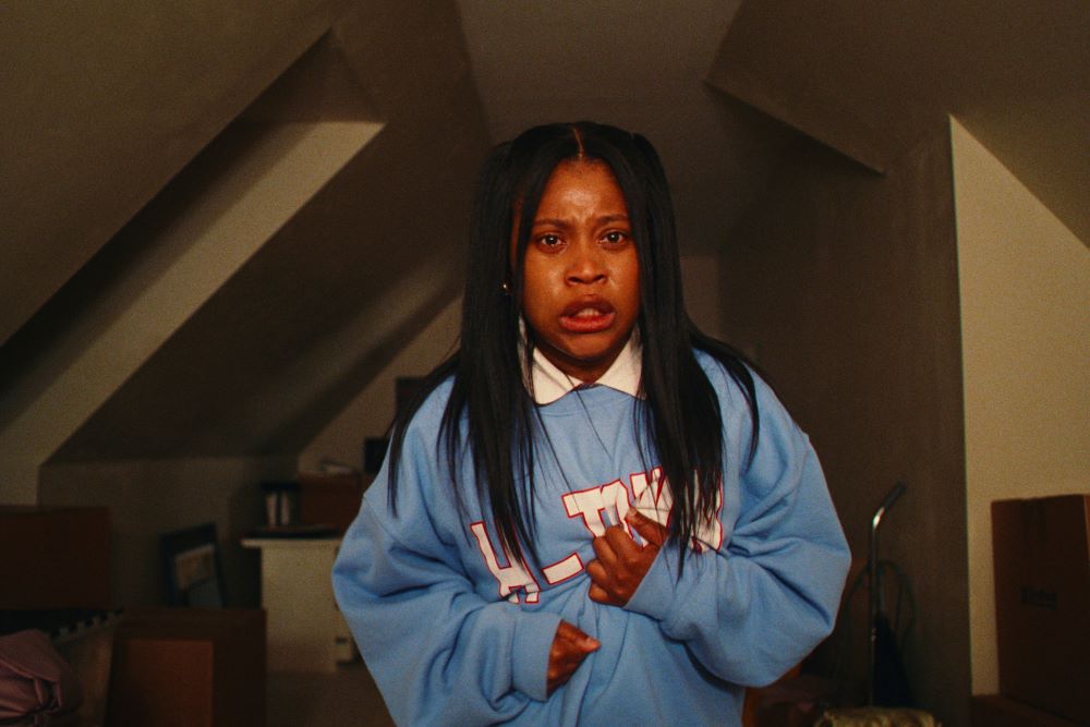 Dominique Fishback as Dre stands in the attic of a house while wearing a powder blue sweatshirt and looking scared in Prime Video's Swarm. 