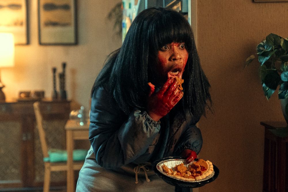 Dre stands in a kitchen with blood smeared on her face and hands while eating pie and looking shocked in Swarm. 