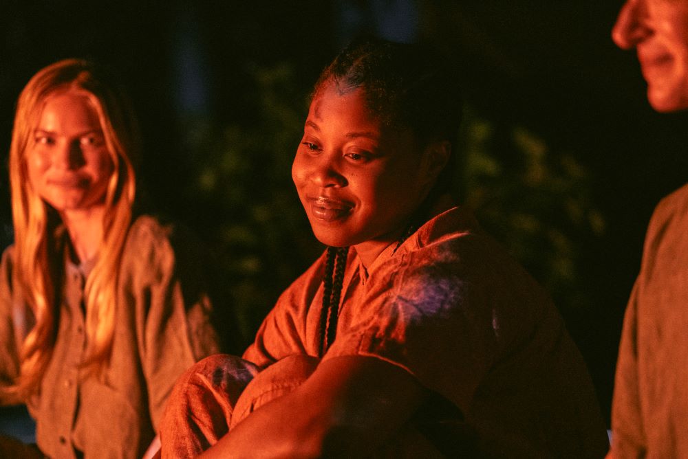 Dominique Fishback as Dre sits around a campfire at night while smiling in Swarm.