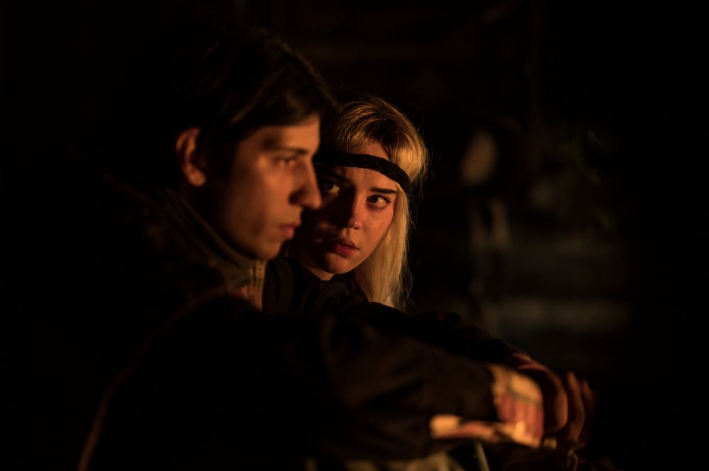 Teen Travis and teen Natalie sit in front of a fire in the cabin.
