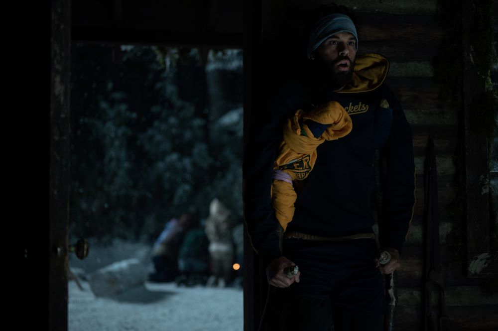 Ben stands in the cabin with the door open, revealing the teens crowded around a fire in the snow.