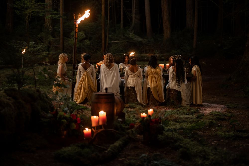 The teens wear Grecian clothing while sitting at a table and surrounded by candles in the wilderness in Yellowjackets Season 2 Episode 2, "Edible Complex."