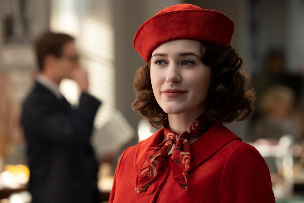 Midge wears a red hat, red jacket and red scarf while smiling and standing in an office in The Marvelous Mrs. Maisel Season 5 Episode 2, "It's a Man, Man, Man, Man World."
