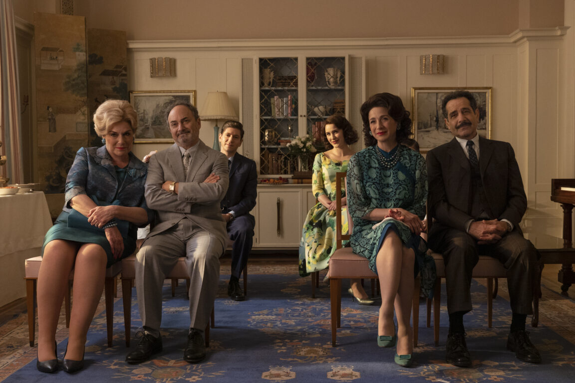 The Maisels and Weissmans sit in the Weissman living room while looking excited in The Marvelous Mrs. Maisel Season 5 Episode 5, "The Pirate Queen."