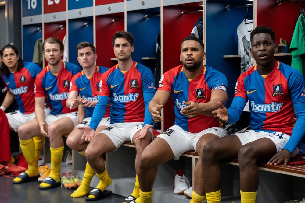 The AFC Richmond football players sit on a bench in their locker room while transfixed by something off screen in Ted Lasso Season 3 Episode 5, "Signs."