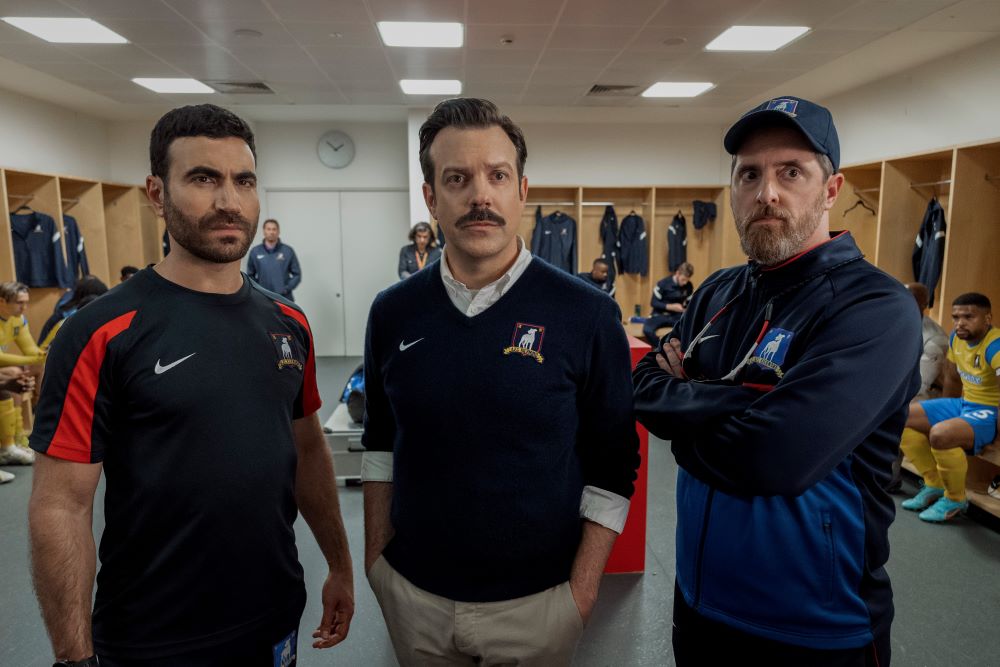 Roy, Ted and Coach Beard stare at something intently while standing in a locker room in Ted Lasso Season 3 Episode 7, "The Strings That Bind Us."