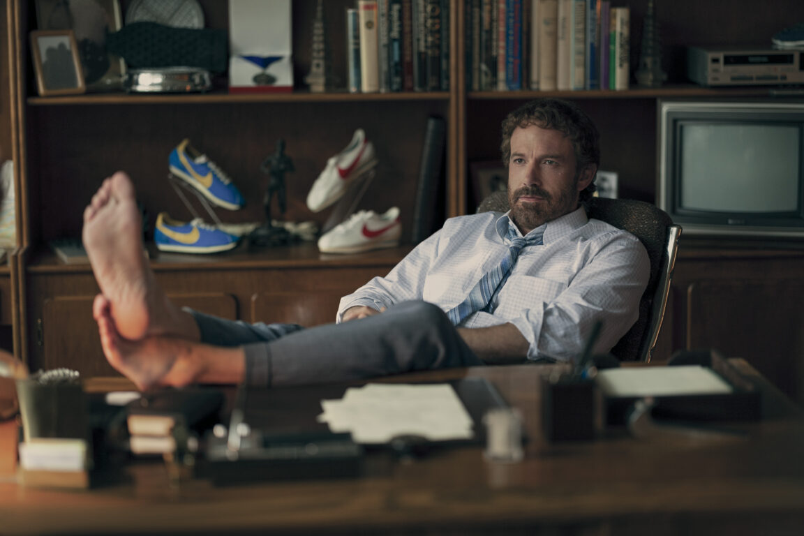 Ben Affleck sits behind his desk with his bare feet kicked up in front of him in the movie Air. 