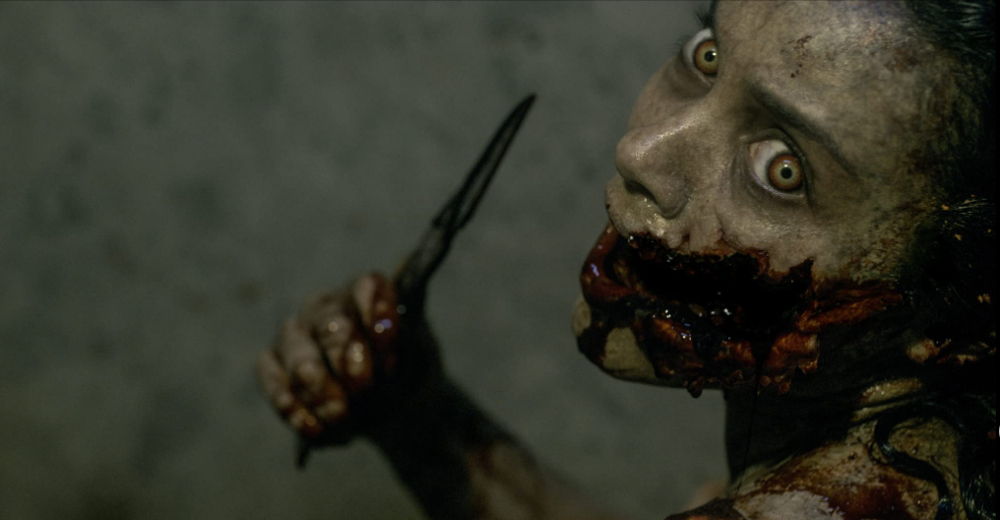 A Deadite holds a knife and looks back at the camera with a bloody face in Evil Dead (2013). 