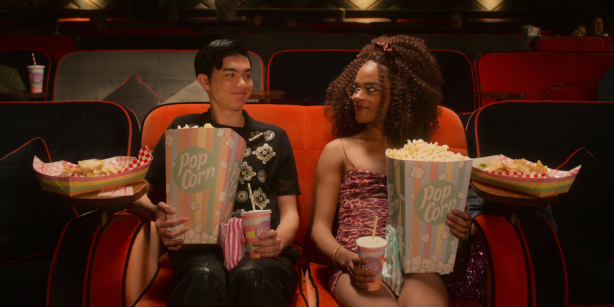 Will Gao aas Tao and Yasmin Finney as Elle in a scene from Heartstopper season 2. The two sit in movie theater seats, looking at each other and surrounded by snacks.