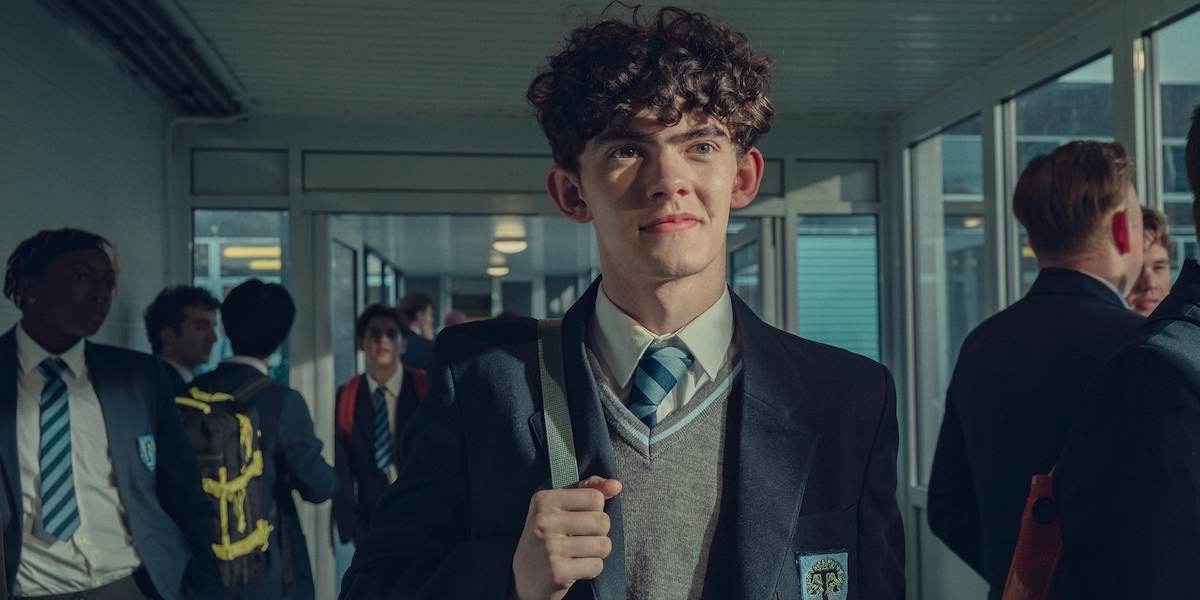 Joe Locke as Charlie Spring in a scene from season two of Heartstopper. He stands in the school hallway surrounded by classmates.