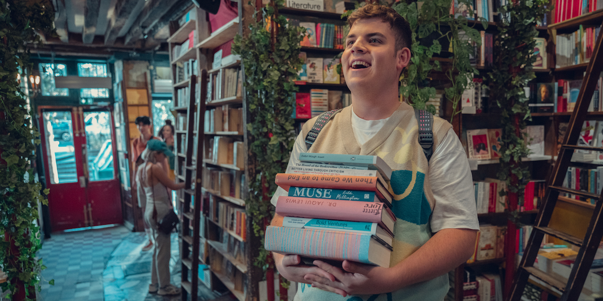 Tobie Donovan as Isaac in a scene from season two of Heartstopper. He's carrying a stack of books in a bookstore full of ladders and plants along the shelves. He has a big smile on his face.
