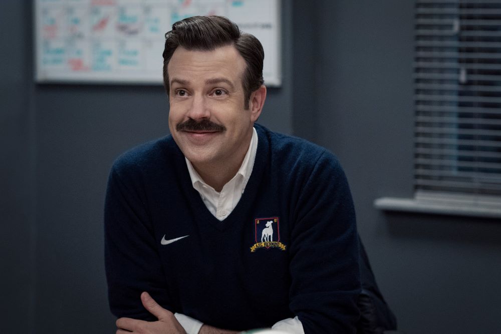 Ted sits at his desk while smiling in Ted Lasso Season 3 Episode 9, "La Locker Room Aux Folles."