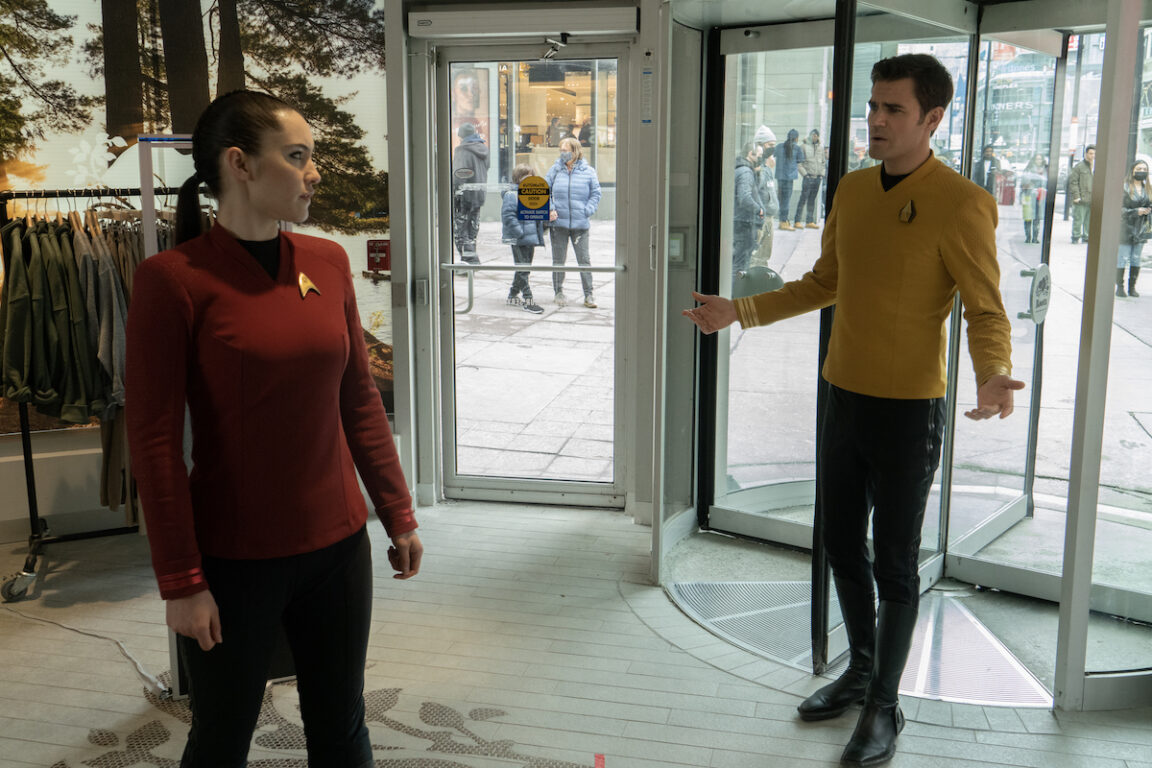 Christina Chong as La’an and Paul Wesley as Kirk in episode 203 “Tomorrow and Tomorrow and Tomorrow” of Star Trek: Strange New Worlds, streaming on Paramount+, 2023. They are both wearing Starfleet uniforms in a 21st century Toronto clothing store.