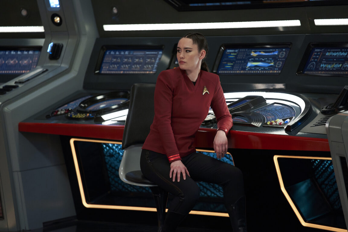 Christina Chong as La'an of the Paramount+ original series STAR TREK: STRANGE NEW WORLDS. She is dressed in her uniform and seated at her station on the bridge of the USS Enterprise. She looks over her shoulder at someone (not pictured).