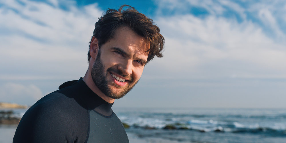 Matt smiles while standing on the beach and wearing a surf suit in Based on a True Story.