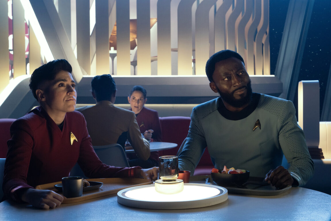 Melissa Navia as Ortegas and Babs Olusanmokun as Dr. M’Benga in episode 202 “Ad Astra per Aspera” of Star Trek: Strange New Worlds, streaming on Paramount+, 2023. They are both seated at a table in the Enterprise cafeteria with food on their trays. They watch Spock converse with the Judge Advocate.