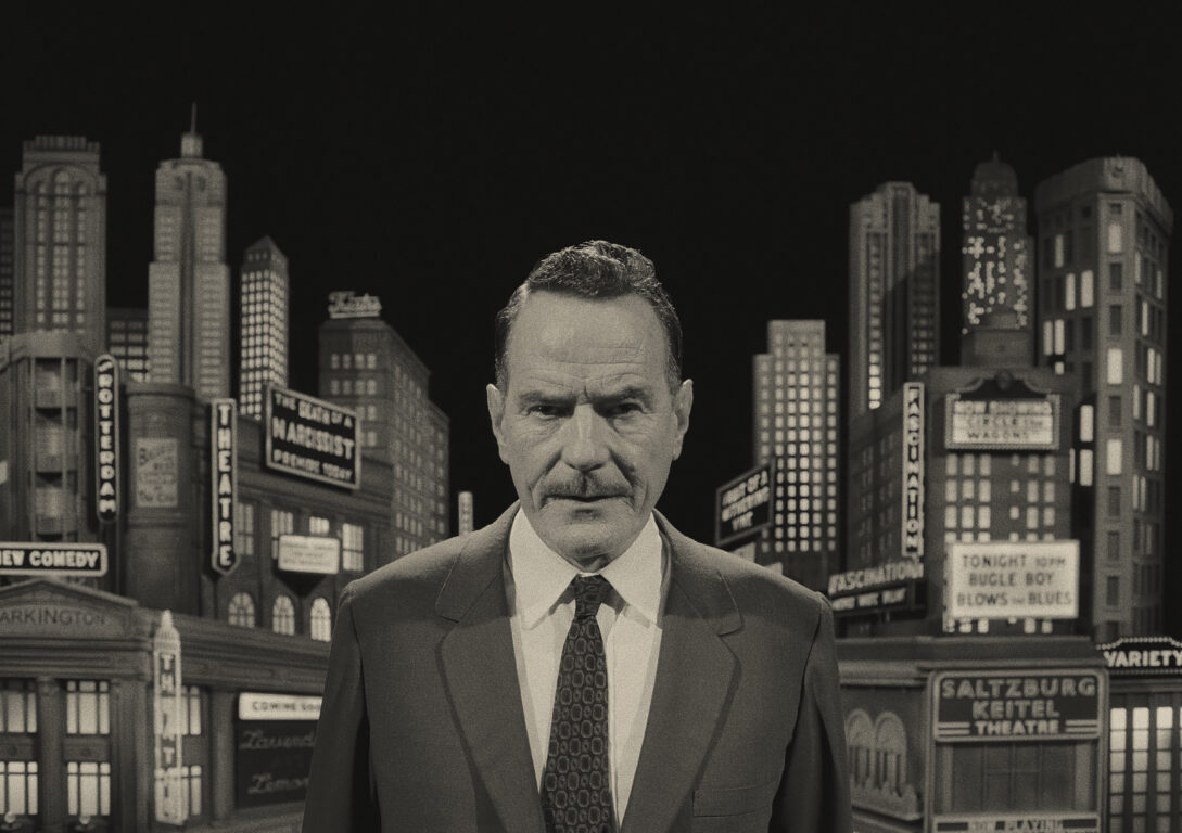 Bryan Cranston smirks at the camera as he stands in front of a New York City artificial backdrop in Asteroid City.