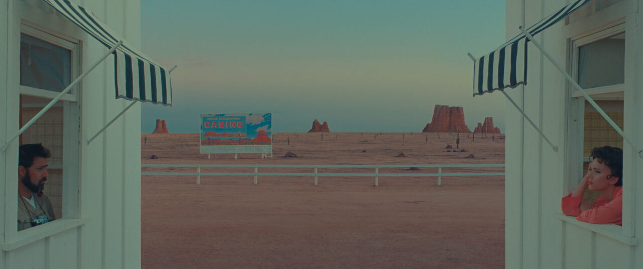 Jason Schwartzman and Scarlett Johansson lean out their motel room windows to talk in front of a desolate desert landscape in Asteroid City.