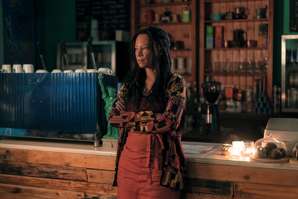 Nina stands in front of the counter in her coffee shop with her arms folded in front of her in Good Omens Season 2 Episode 6, "Chapter 6: Everyday."