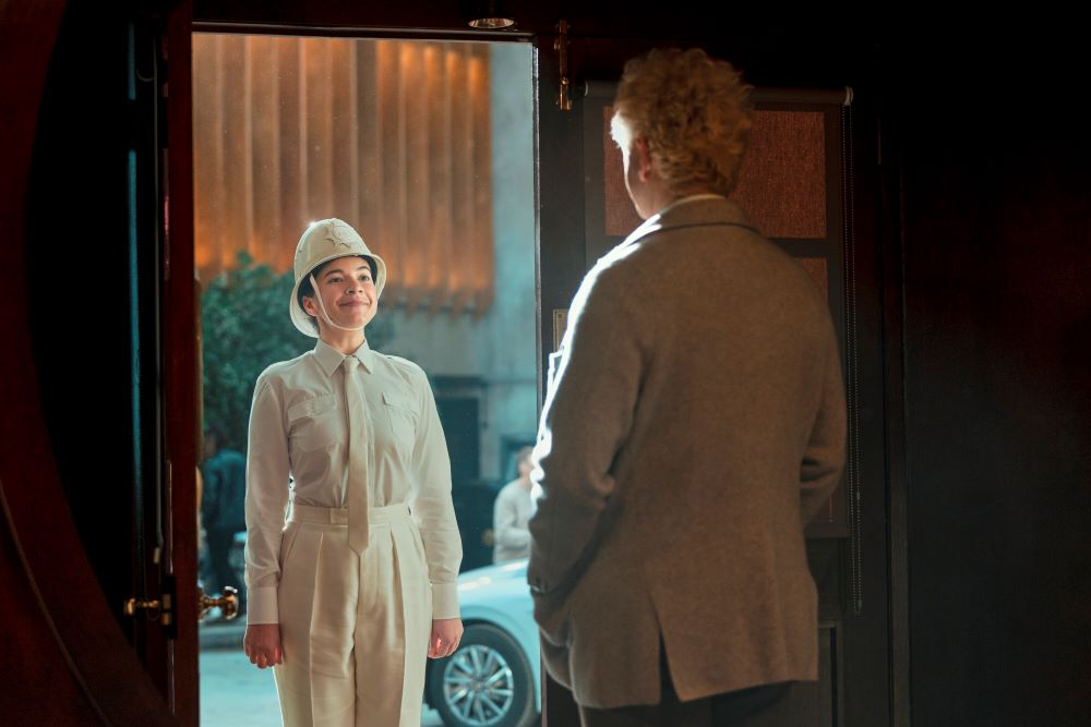 Muriel wears a white constable's uniform while standing on Aziraphale's bookstore doorstep and smiling in Good Omens Season 2 Episode 3, "Chapter 3: I Know Where I'm Going."