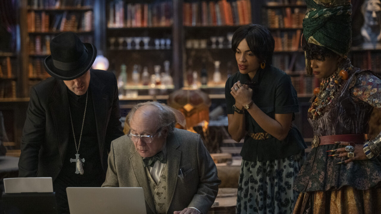 Owen Wilson, Danny DeVito, Rosario Dawson and Tiffany Hadish look over a laptop screen as they stand in a ornate room.