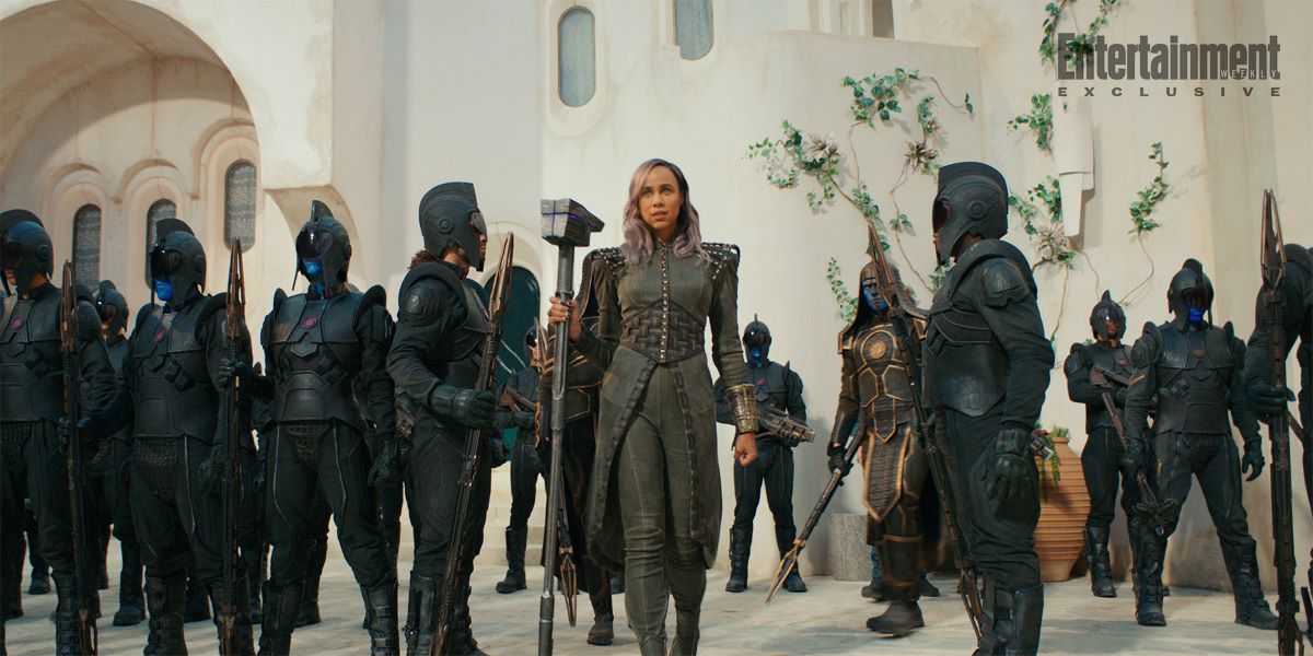 Dar-Benn wears an all-gray ensemble with a long coat and boots while holding a gray scepter and surrounded by her guards.