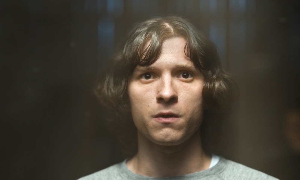 A white man (Tom Holland) in his 20s with medium-length brown hair stares almost into the camera. The lighting is bright, but the background is dark.