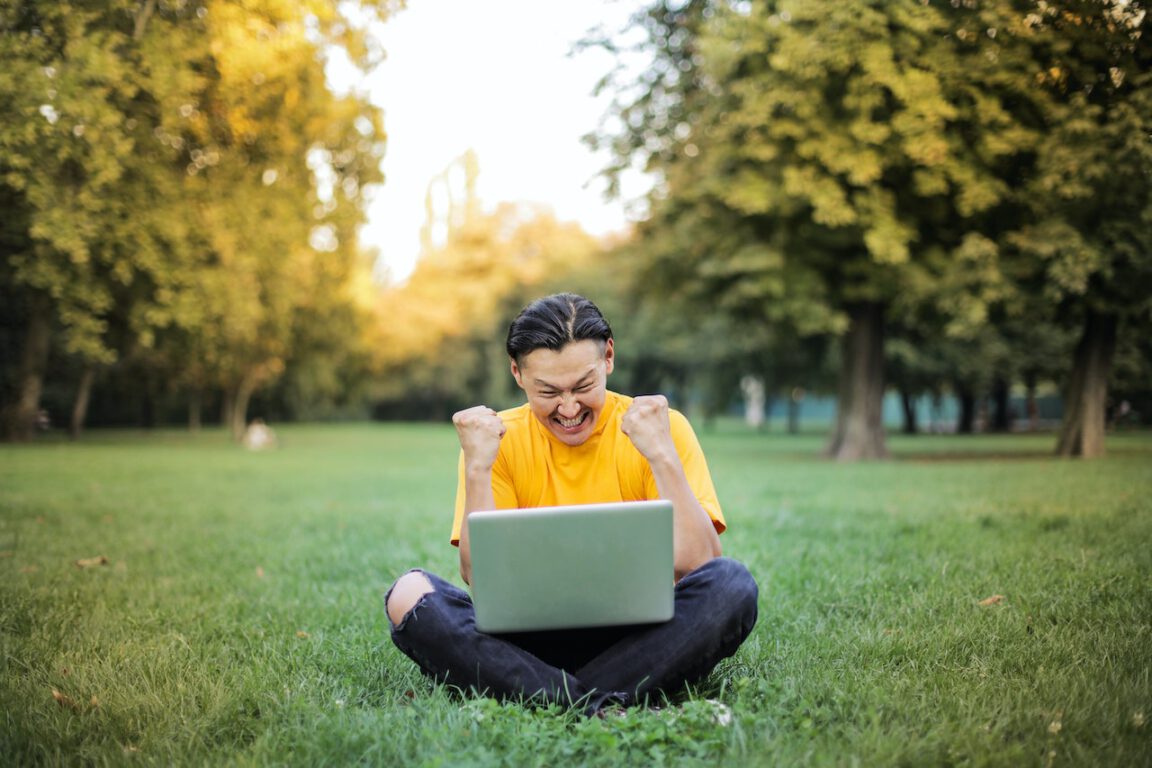 a man with dark pants and a yellow shirt sits on grass in a park. His fists are raised in triumph as he looks at his laptop screen.