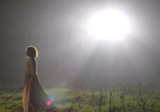 A woman walks through a field with a bright light shinning down from the sky in The Village.