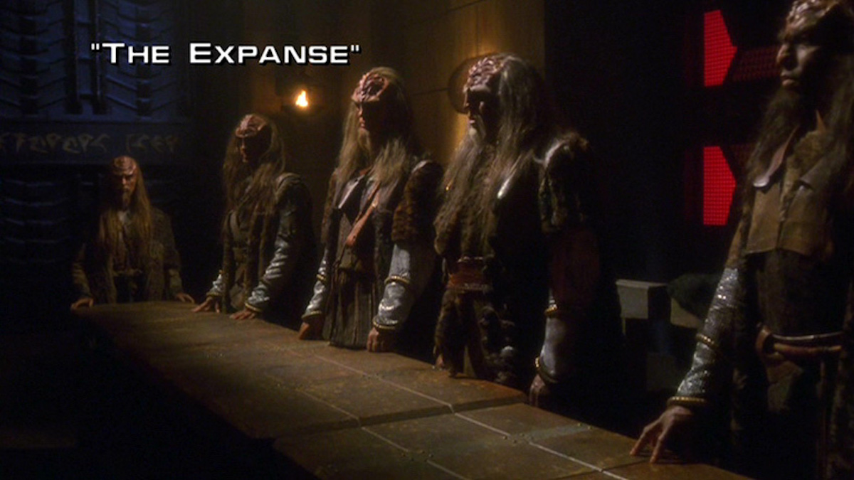 A line of Klingons stand before a table in the title card for Enterprise season 2's "The Expanse."