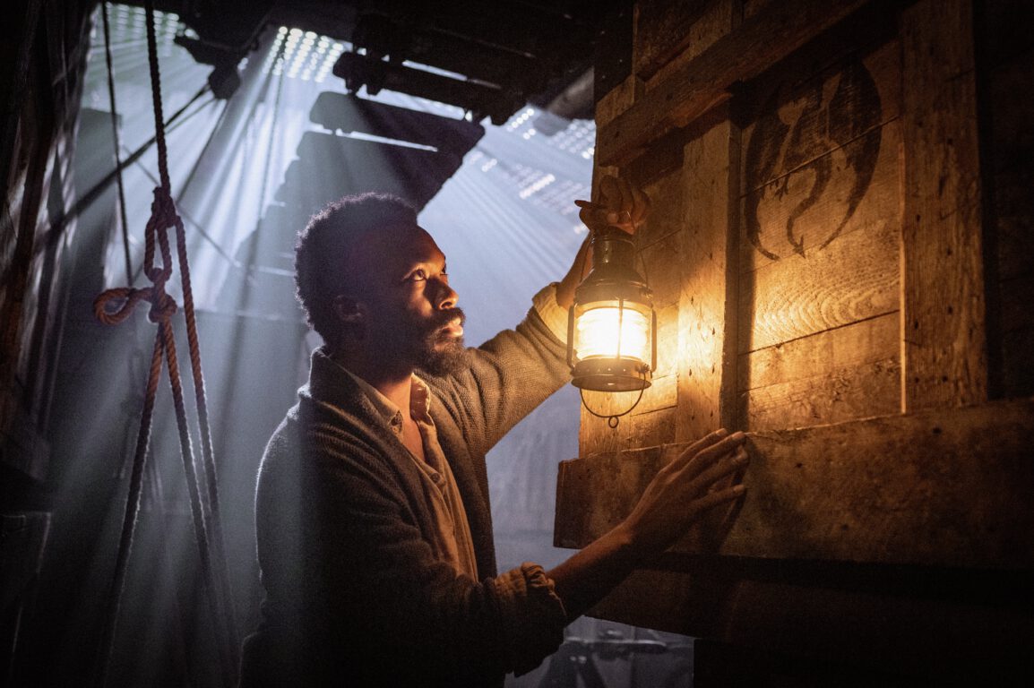 Corey Hawkins holds a lantern and stares at a box with a large dragon logo in a dusty cargo hold.