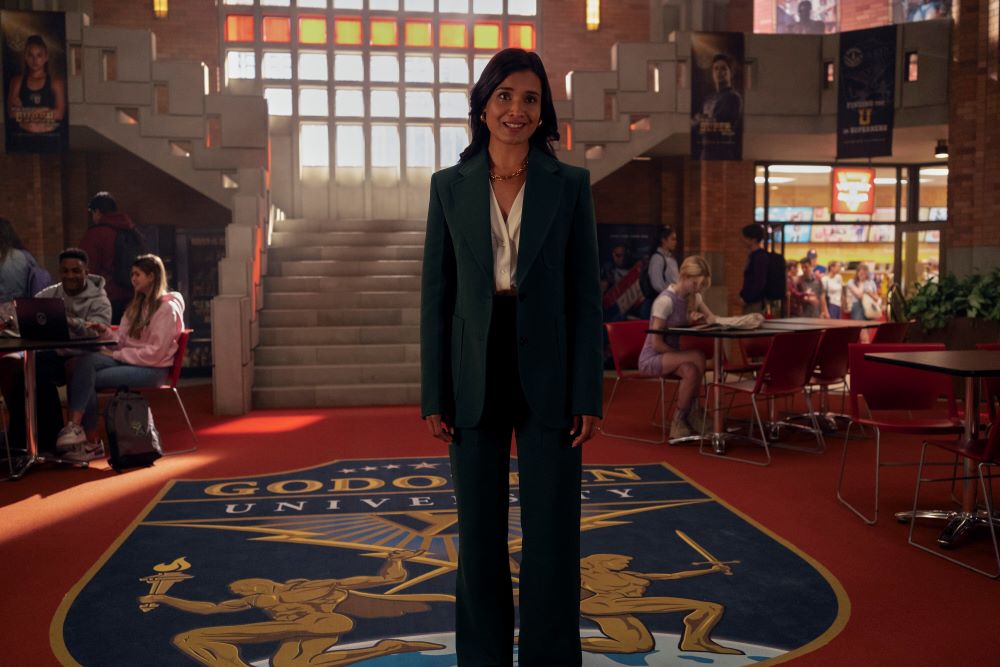 Dean Indira Shetty wears a black blazer with matching pants and a white blouse while standing in a college building and looking forward in Prime Video's Gen V Season 1 Episode 1, "God U."