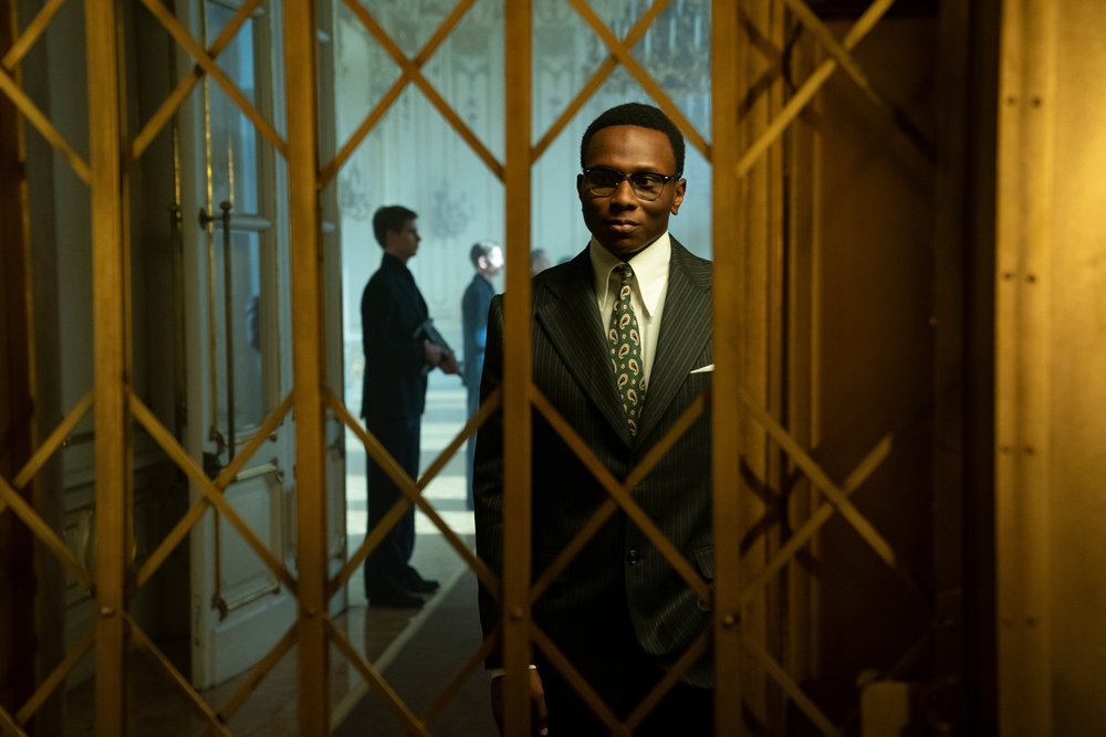 Charon wears a dark suit while standing in front of a gold elevator gate in The Continental: From the World of John Wick, Season 1 Episode 1, "Night 1."