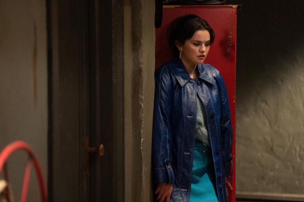 Mabel wears a blue leather jacket while standing in the backstage hallway of a theater and looking nervous in Only Murders in the Building Season 3 Episode 6, "Ghost Light."