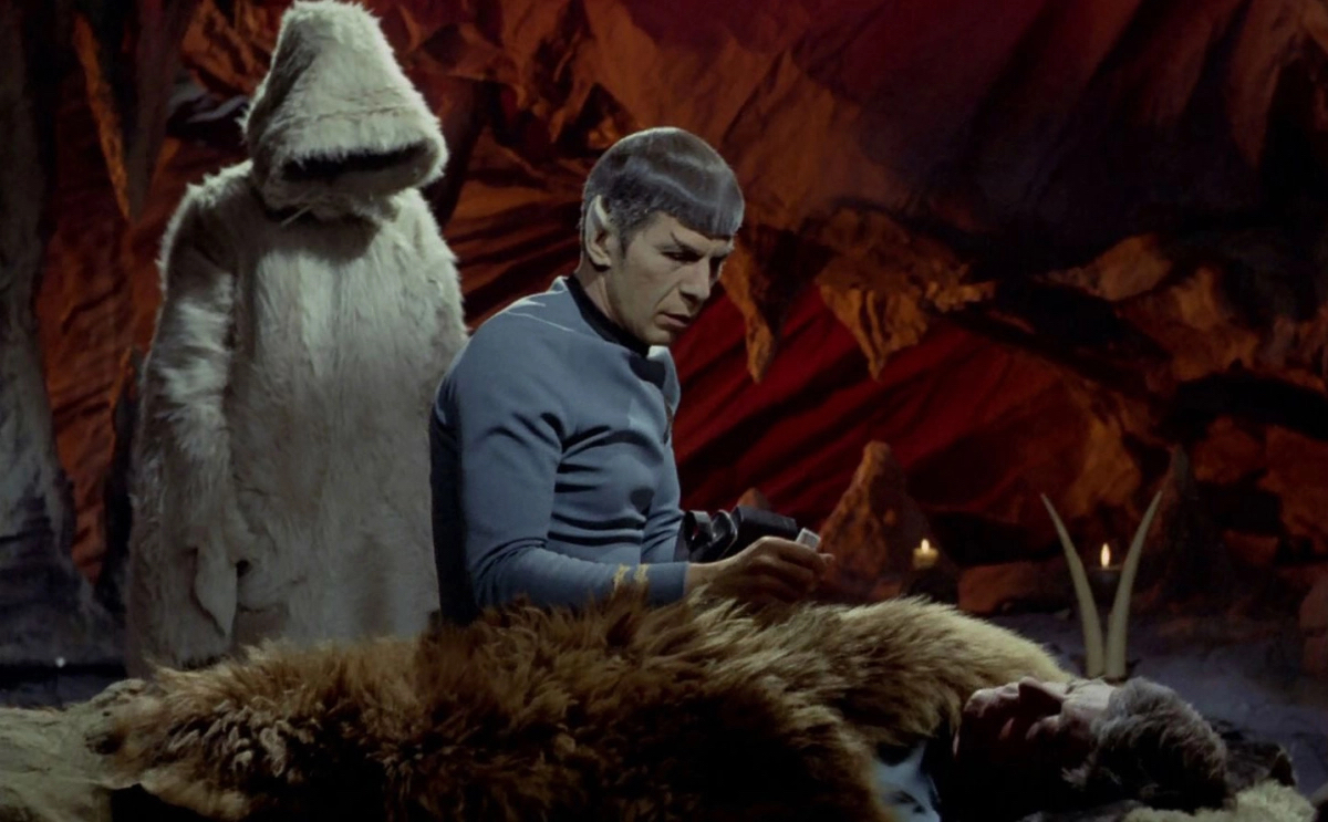 Spock sits near a man who's lying under a fur blanket in a cave while a hooded figure stands above him in Star Trek: The Original Series. 