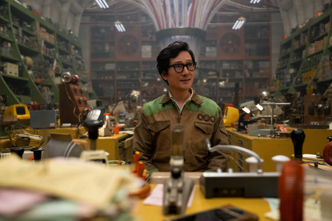 O.B. stands behind the counter in the Repairs & Advancements Department of the TVA while looking content in Loki Season 2 Episode 1, "Ouroboros."