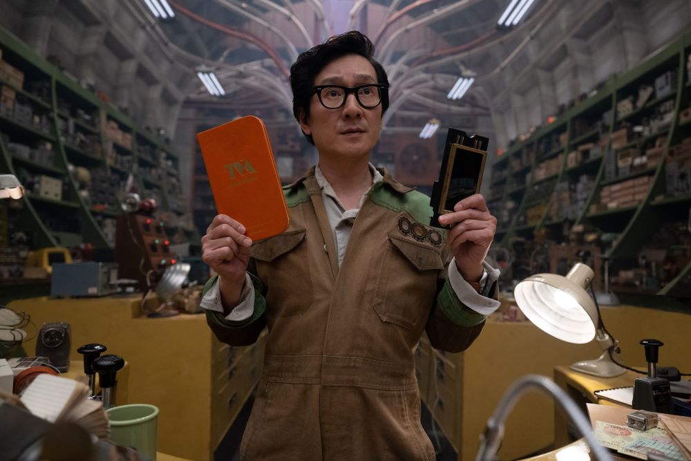 O.B. holds a TVA Guidebook in one hand and a TemPad in another while standing behind a desk and looking serious in Loki Season 2 Episode 2, "Breaking Brad."