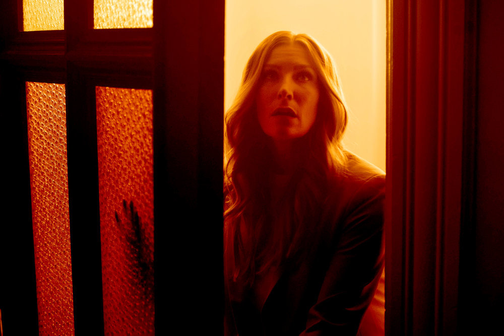 Susan Ireland opens the door to a house while cast in an orangish light in SurrealEstate Season 2 Episode 3, "The Butler Didn't."