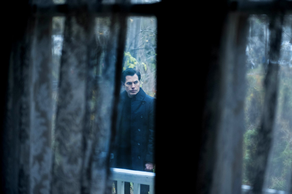 Luke stands outside during the day while someone watches him through a curtained window in their darkened home in SurrealEstate Season 2 Episode 4, "I Put a Spell on You."